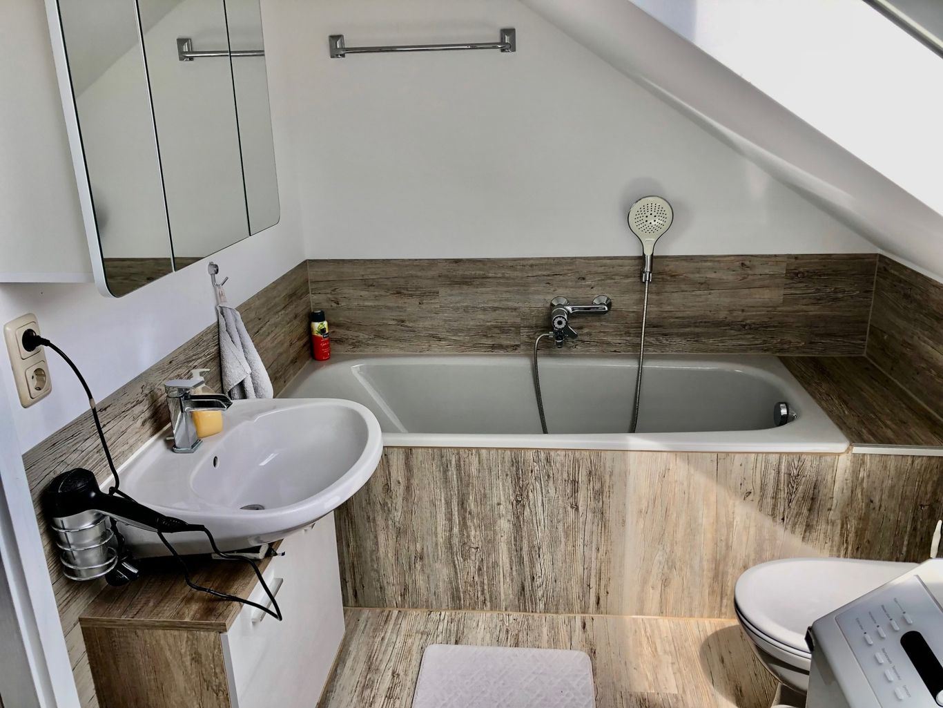 Modernes Badezimmer mit Badewanne, Waschbecken und Spiegel, gestaltet in warmen Holztönen. Eine Dachschräge mit Fenster.