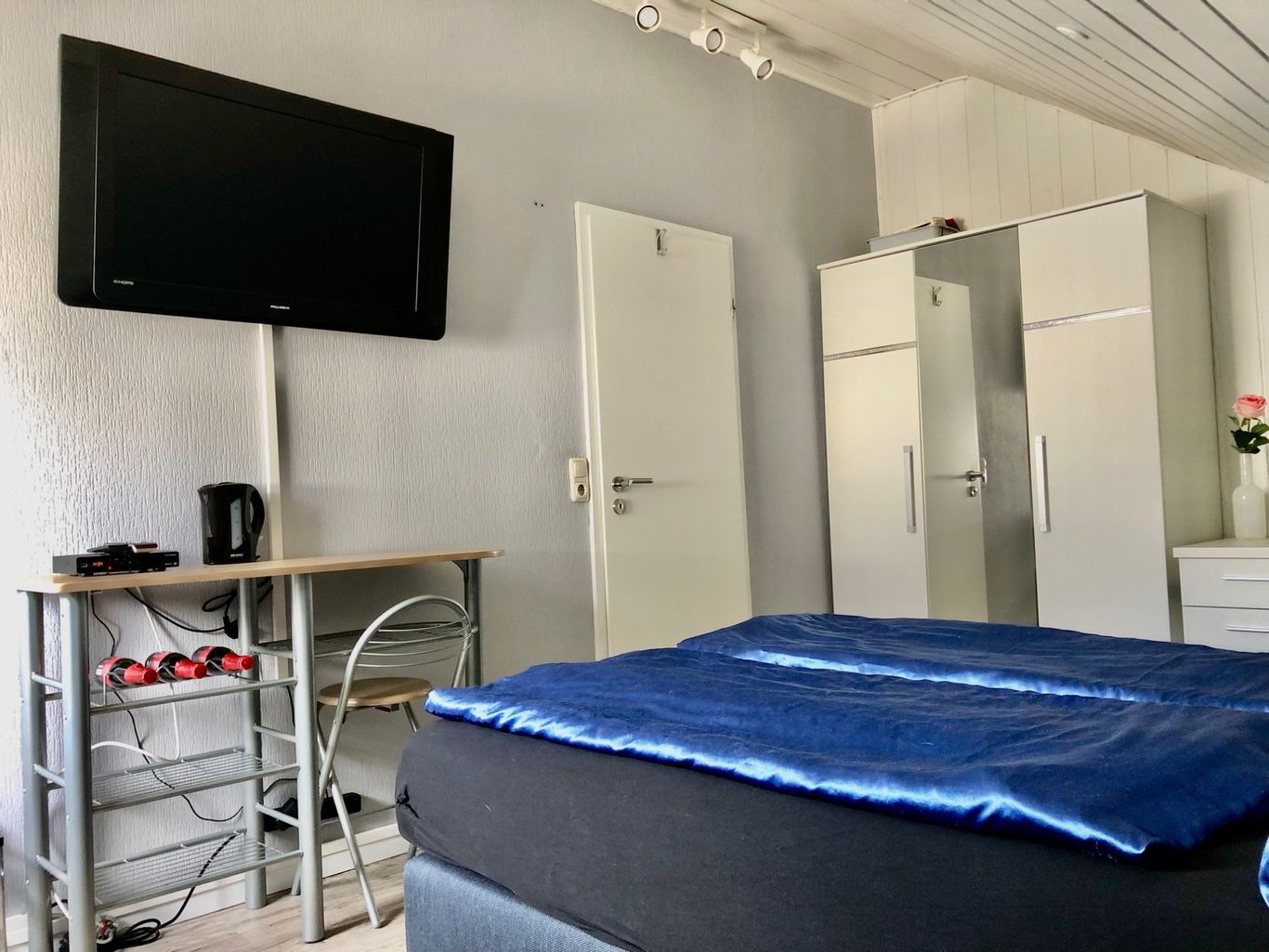 Gemütliches Gästezimmer mit einem großen Bett, Wand-TV, Kleiderschrank und einem kleinen Tisch mit Wasserkocher. 