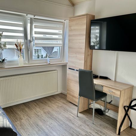 Gemütlicher Arbeitsbereich in einem hellen Zimmer mit großem Fenster, Schreibtisch und Stuhl. Ein Wandschrank bietet zusätzlichen Stauraum.