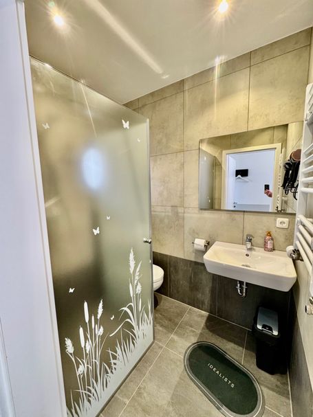Elegantes Badezimmer mit großer, dekorativ verzierter Glasdusche, stilvollem Waschbecken und beleuchtetem Spiegel. 