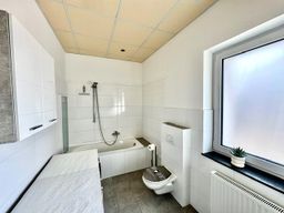 Freundliches Badezimmer mit weiß gefliesten Wänden, Badewanne mit Duschfunktion, WC und Fenster mit Milchglas für Privatsphäre. 